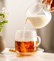 Seitliche Ansicht einer Untertasse mit einer Tasse Tee, in die Milch gegossen wird, neben einer kleinen Blume in einer Vase.