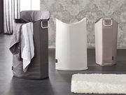 Drei Wäschesammler in verschiedenen Farben, von links nach rechts: dunkelgrau mit Handtuch, weiß und beige, auf einem dunklen Boden vor einer gemusterten Wand, Perspektive von vorne.