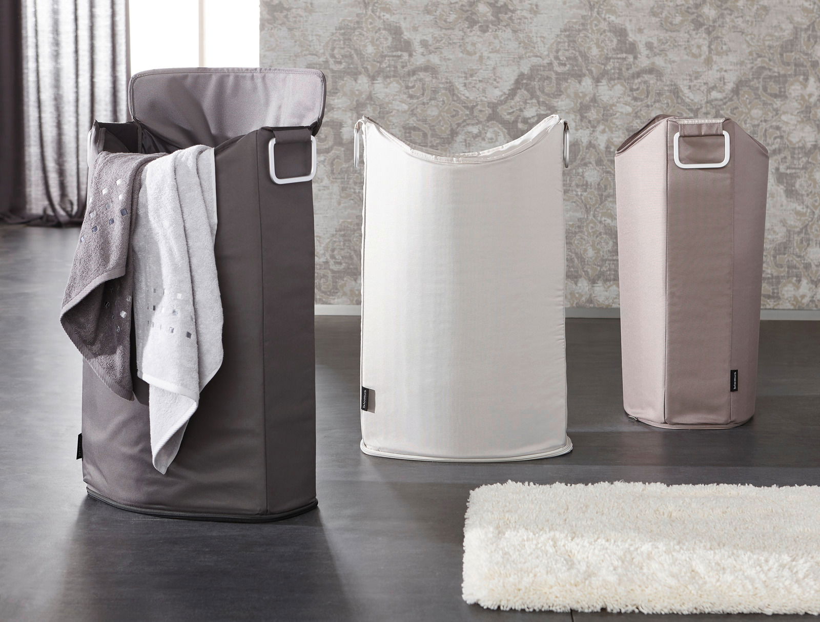 Drei Wäschesammler in verschiedenen Farben, von links nach rechts: dunkelgrau mit Handtuch, weiß und beige, auf einem dunklen Boden vor einer gemusterten Wand, Perspektive von vorne.