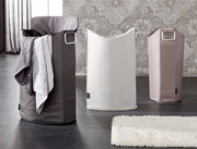 Drei Wäschesammler in verschiedenen Farben, von links nach rechts: dunkelgrau mit Handtuch, weiß und beige, auf einem dunklen Boden vor einer gemusterten Wand, Perspektive von vorne.