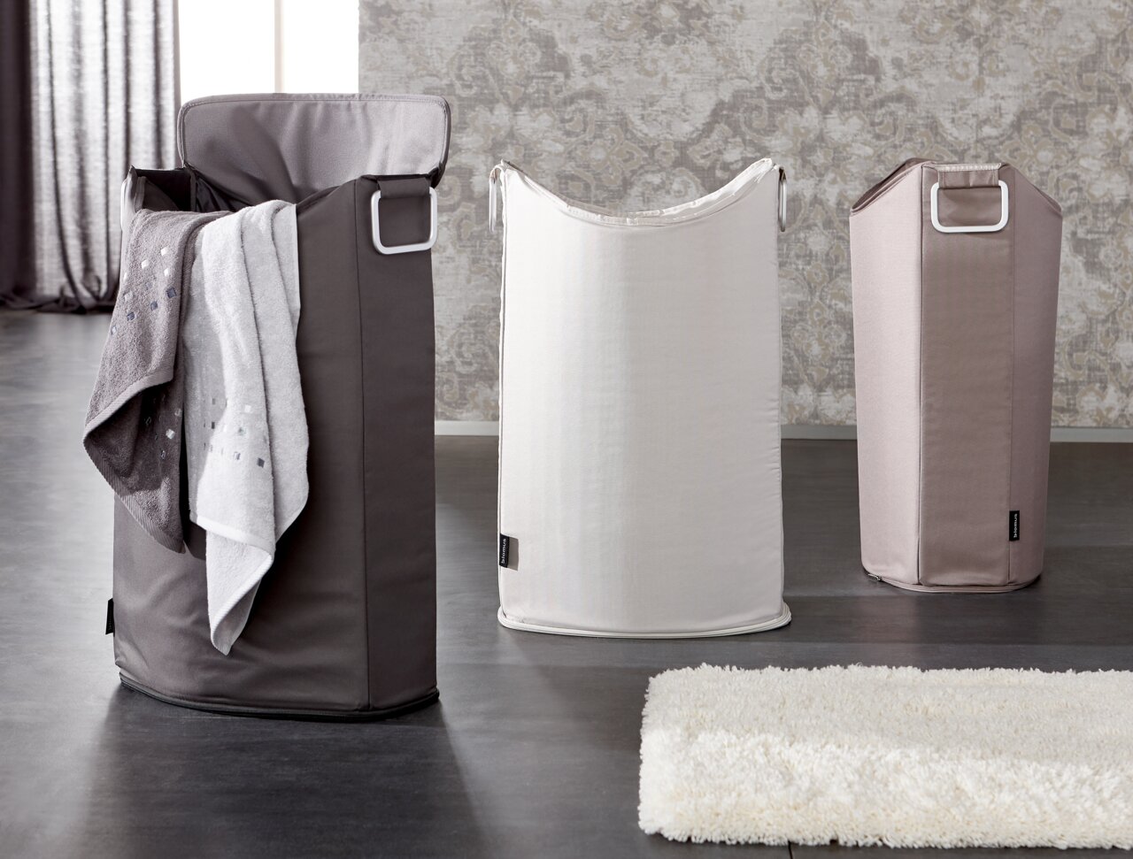 Drei Wäschesammler in verschiedenen Farben, von links nach rechts: dunkelgrau mit Handtuch, weiß und beige, auf einem dunklen Boden vor einer gemusterten Wand, Perspektive von vorne.