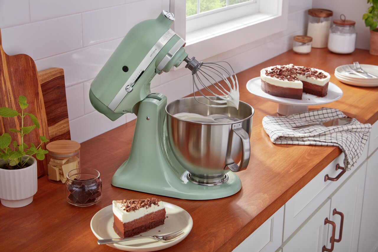 Küchenmaschine Artisan in mintgrün auf einer Küchenarbeitsplatte, seitliche Perspektive, mit Rührbesen und Rührschüssel, umgeben von Kuchen und Zutaten.
