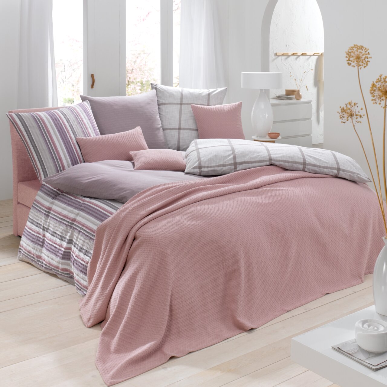 Linneweber Sprei SUMMER Een gezellig slaapkamerbeeld met een roze sprei op het bed, omringd door sierkussens in verschillende patronen en kleuren.