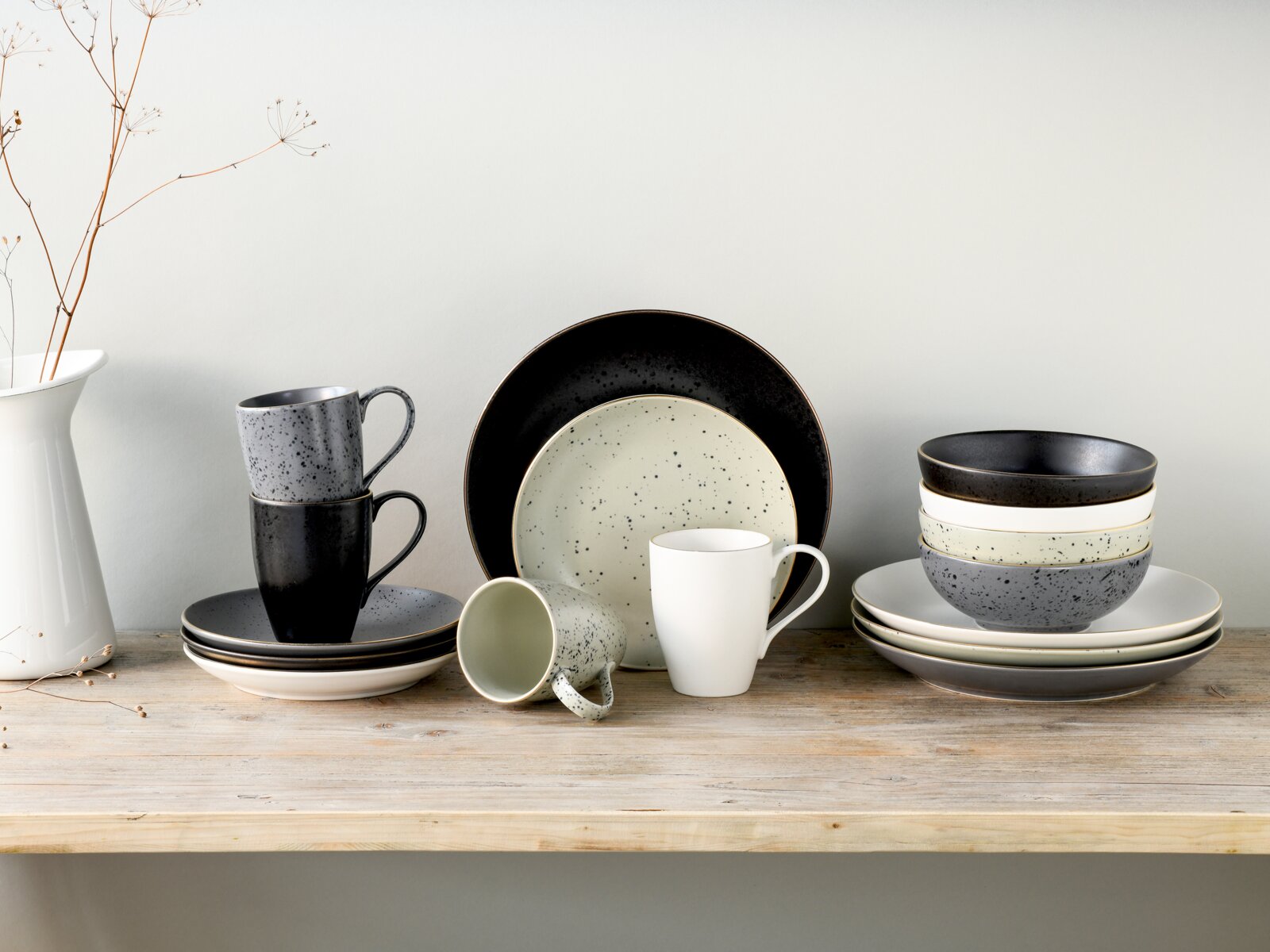 creaTable Kombiservice 16-tlg INDUSTRIAL Modernes, gesprenkeltes Keramik-Geschirrset in Schwarz, Grau und Creme mit Tellern, Schalen und Tassen auf Holzbrett vor heller Wand, seitliche Frontansicht auf Augenhöhe