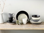 Modernes, gesprenkeltes Keramik-Geschirrset in Schwarz, Grau und Creme mit Tellern, Schalen und Tassen auf Holzbrett vor heller Wand, seitliche Frontansicht auf Augenhöhe