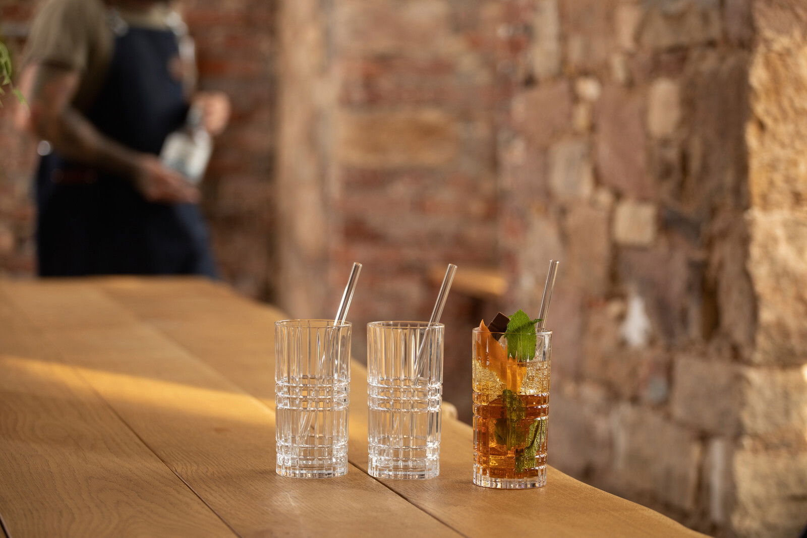 Drei Longdrinkgläser mit Strohhalmen auf einem Holztisch, eines gefüllt mit einem Getränk und Garnitur, seitliche Perspektive.