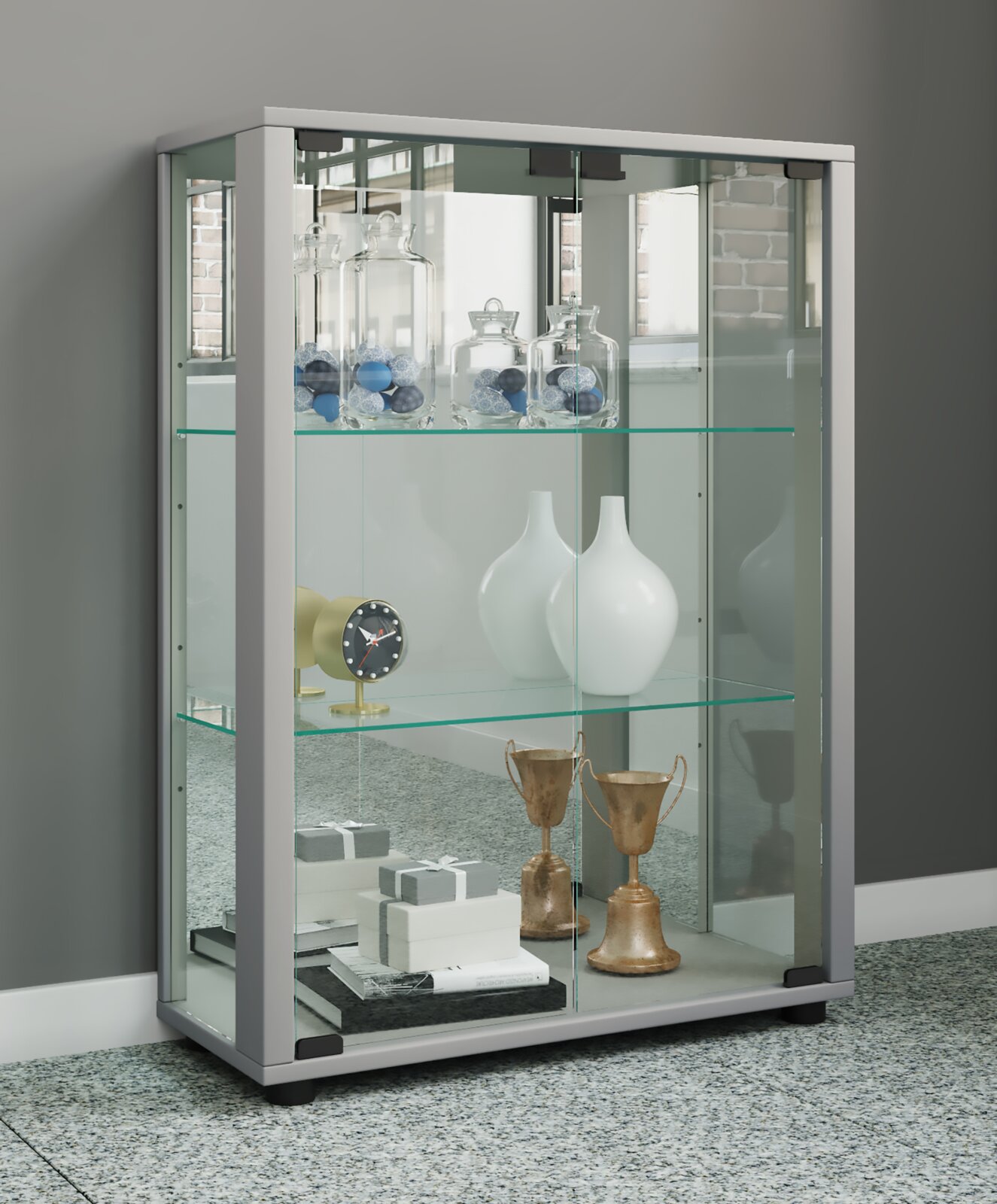 Moderne Glasvitrine mit weiß-grauem Rahmen und Glasböden, befüllt mit Dekogläsern, Vasen und Pokalen; perspektivische Ansicht schräg von vorn links.