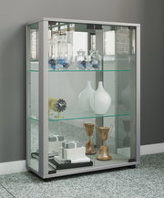 Moderne Glasvitrine mit weiß-grauem Rahmen und Glasböden, befüllt mit Dekogläsern, Vasen und Pokalen; perspektivische Ansicht schräg von vorn links.