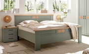 Modernes Doppelbett in Salbeigrün mit Holzauflagen und dekorativen Holzeinsätzen, im hellen Schlafzimmer mit Nachttisch und Kleiderschrank, fotografiert aus schräger Frontansicht.
