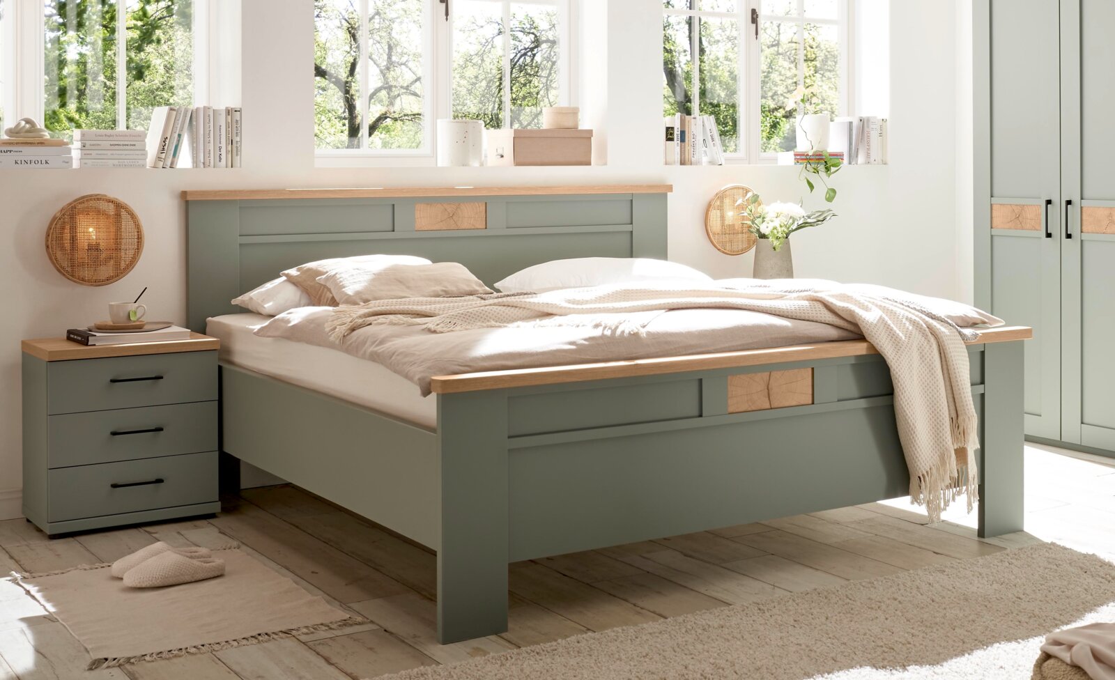 Modernes Doppelbett in Salbeigrün mit Holzauflagen und dekorativen Holzeinsätzen, im hellen Schlafzimmer mit Nachttisch und Kleiderschrank, fotografiert aus schräger Frontansicht.