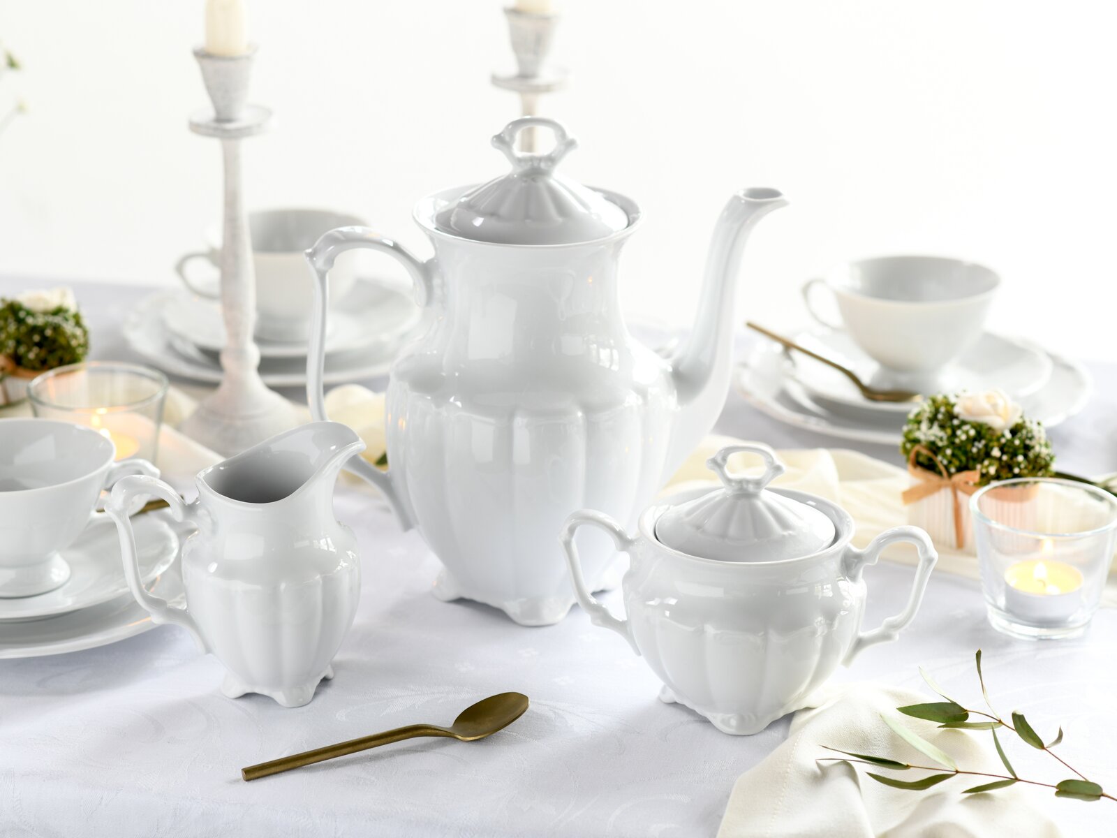 Weißes Porzellan-Kaffeeservice mit Kaffeekanne, Zuckerdose und Milchkännchen auf gedecktem Tisch, leicht von vorne in Augenhöhe fotografiert.