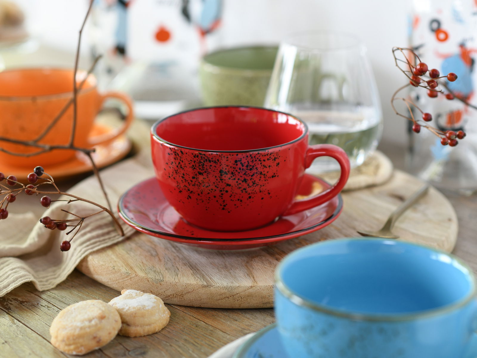 Rote Cappuccinotasse mit Untertasse, umgeben von weiteren bunten Tassen und Dekorationen auf einem Holztisch, aus einer leicht seitlichen Perspektive fotografiert.