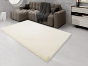 Ein cremefarbener Fellteppich liegt in einem modernen Wohnzimmer vor einem grauen Sofa. Die Perspektive zeigt den Teppich von schräg oben.