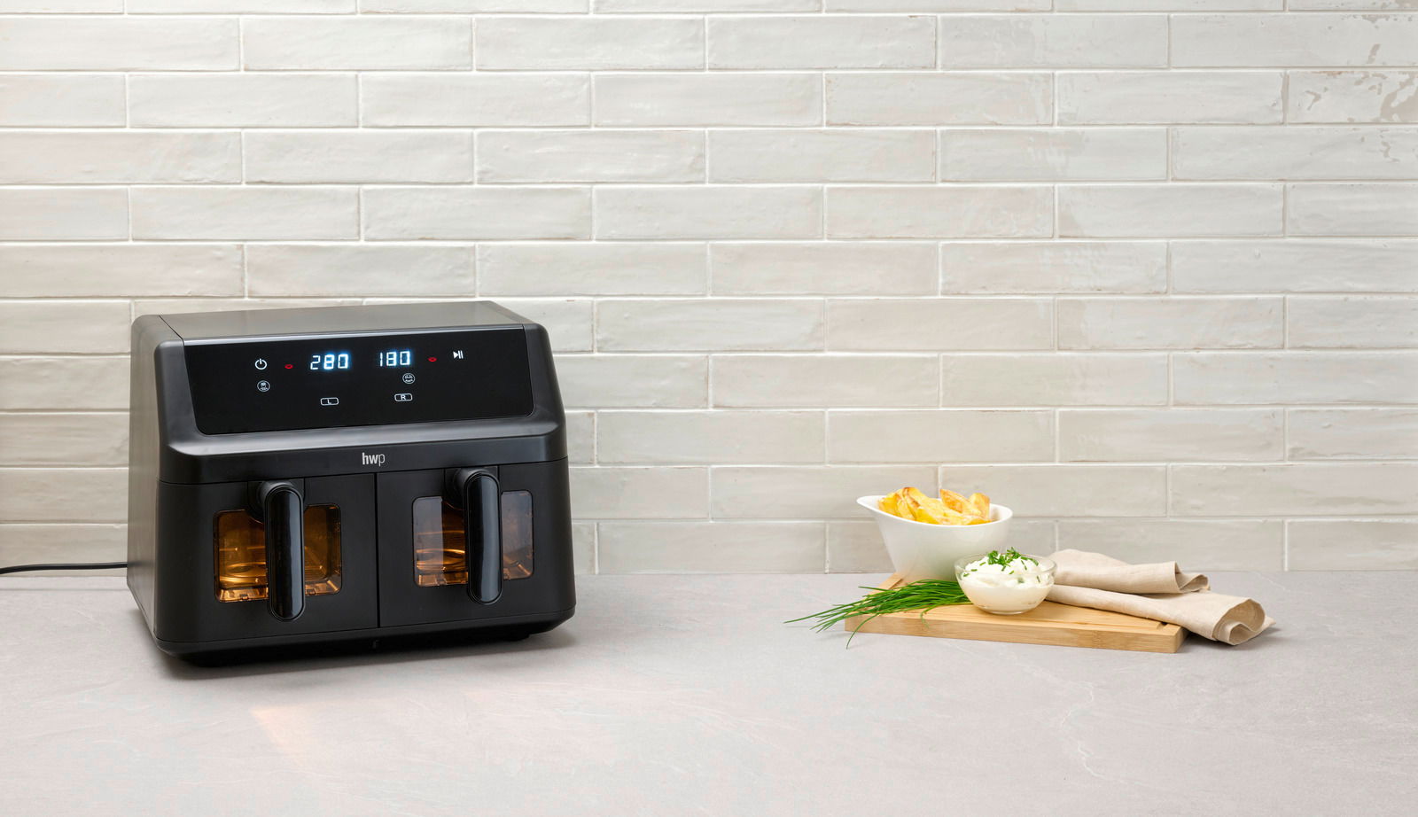 Schwarze Heißluftfritteuse mit digitalem Display, frontal auf einer Küchenarbeitsplatte platziert, daneben eine Schale mit Pommes und Dip.