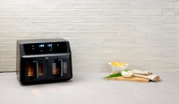 Schwarze Heißluftfritteuse mit digitalem Display, frontal auf einer Küchenarbeitsplatte platziert, daneben eine Schale mit Pommes und Dip.