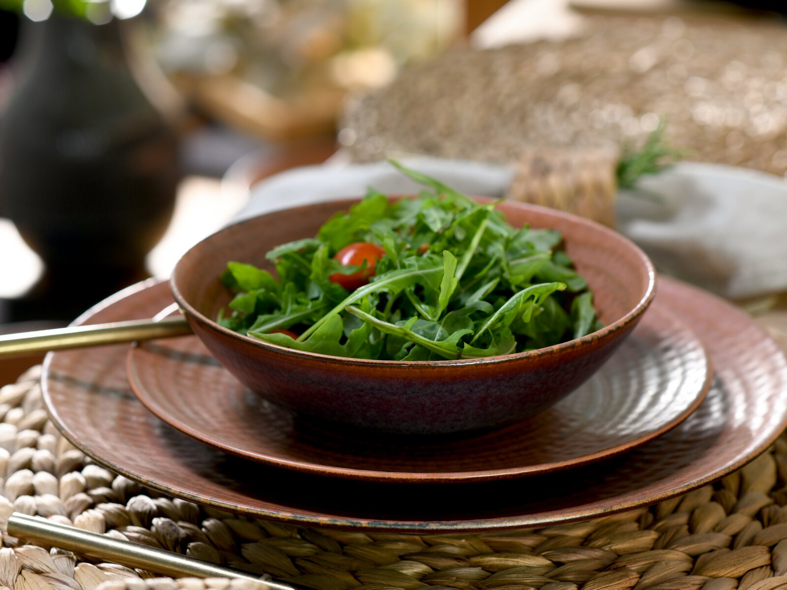 Braune Keramik-Schüssel mit frischem Rucola- und Tomatensalat auf passendem Teller, seitliche Nahaufnahme in leicht erhöhter Perspektive auf einem gedeckten Tisch