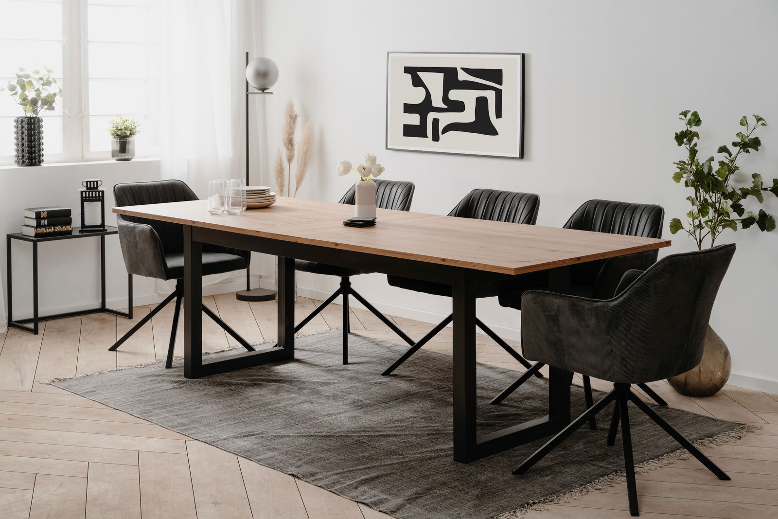 Moderner Esstisch mit Holzplatte und schwarzen Metallbeinen, umgeben von schwarzen Stühlen in einem hellen Raum mit Kunst an der Wand, aus seitlicher Perspektive.