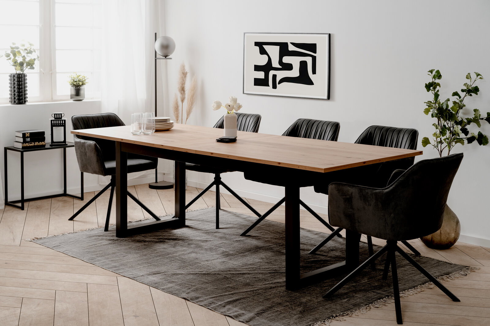 Moderner Esstisch mit Holzplatte und schwarzen Metallbeinen, umgeben von schwarzen Stühlen in einem hellen Raum mit Kunst an der Wand, aus seitlicher Perspektive.