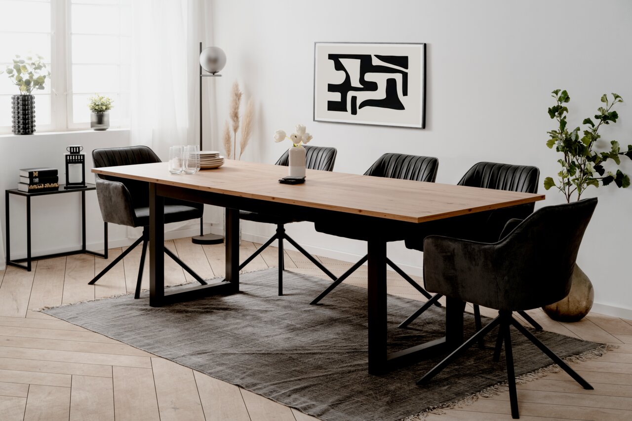 Moderner Esstisch mit Holzplatte und schwarzen Metallbeinen, umgeben von schwarzen Stühlen in einem hellen Raum mit Kunst an der Wand, aus seitlicher Perspektive.