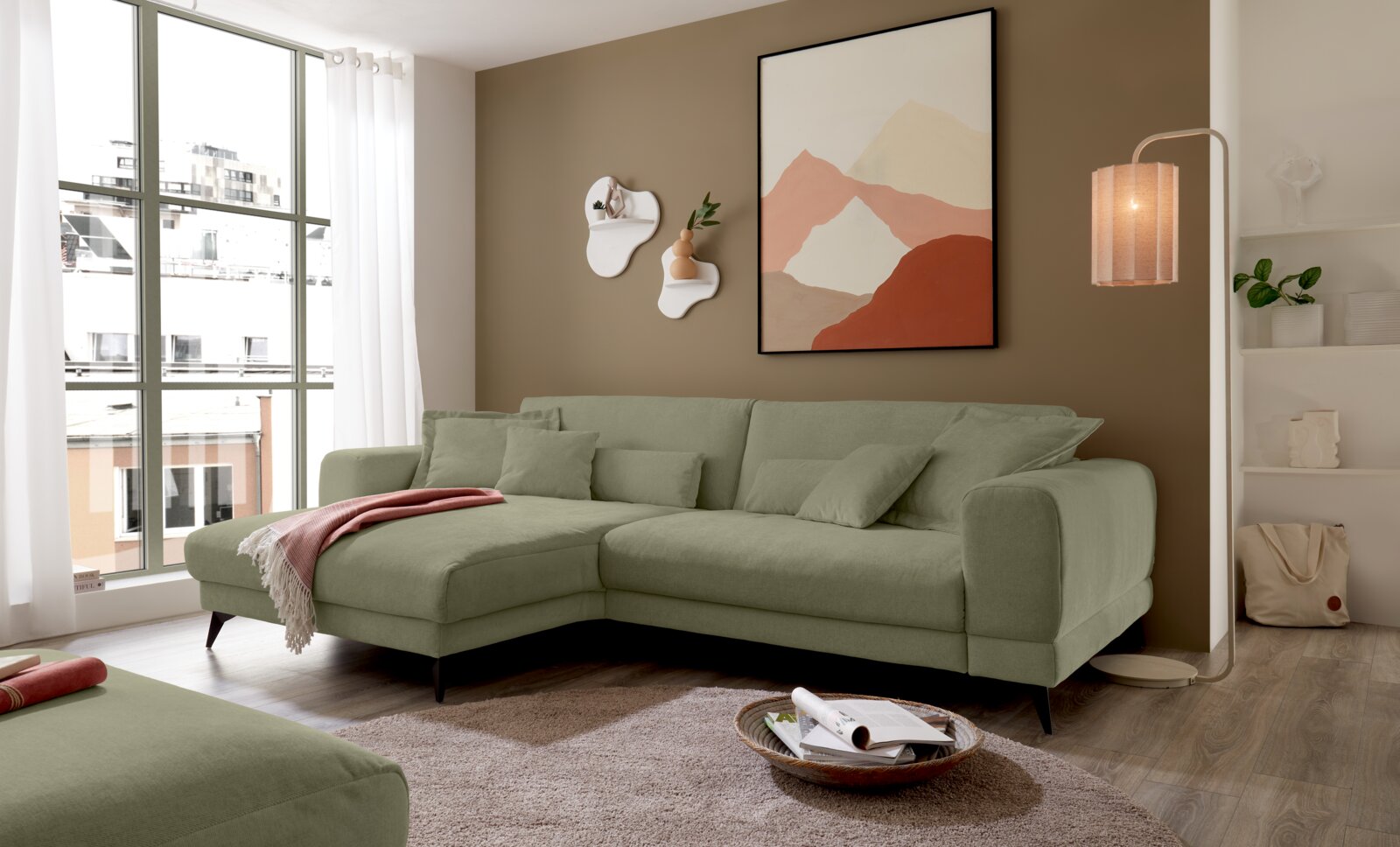 Set one by Musterring Ecksofa SET ONE MIDLAND Modernes grünes L-förmiges Ecksofa mit Ottomane links und schwarzen Füßen im Wohnzimmer, Frontansicht leicht schräg von links, dekoriert mit Kissen und Decke, daneben Stehlampe und Wandbild.