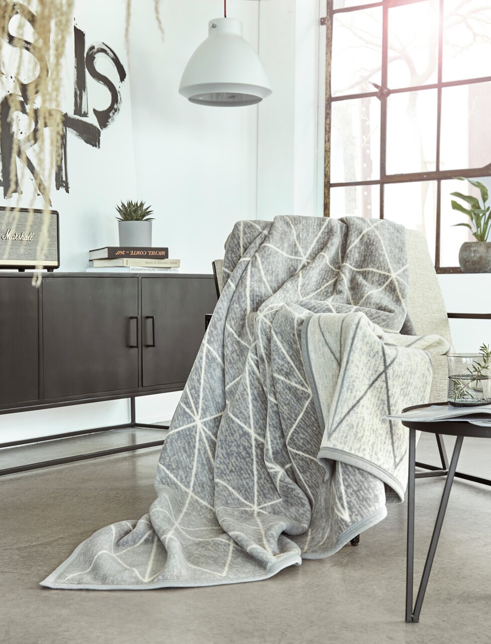 IBENA Wohndecke 150x200cm BUGATTI EX Gemütliche Jacquard Decke in modernem geometrischen Muster, drapiert über einem Sessel in einem stilvollen Wohnzimmer mit Blick auf ein großes Fenster.