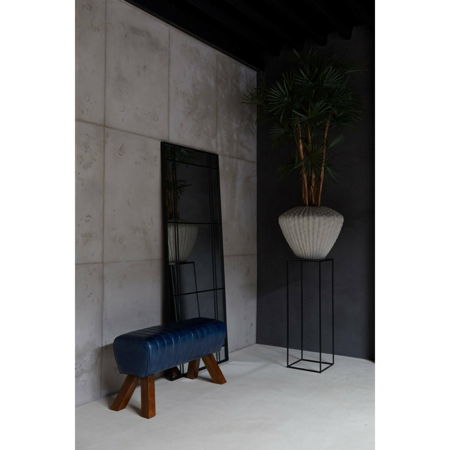 Großer rechteckiger Spiegel mit schwarzem Rahmen, der an einer Wand lehnt, neben einer blauen Bank und einer hohen Pflanze in einem modernen Raum. Perspektive von der Seite.