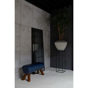 Großer rechteckiger Spiegel mit schwarzem Rahmen, der an einer Wand lehnt, neben einer blauen Bank und einer hohen Pflanze in einem modernen Raum. Perspektive von der Seite.