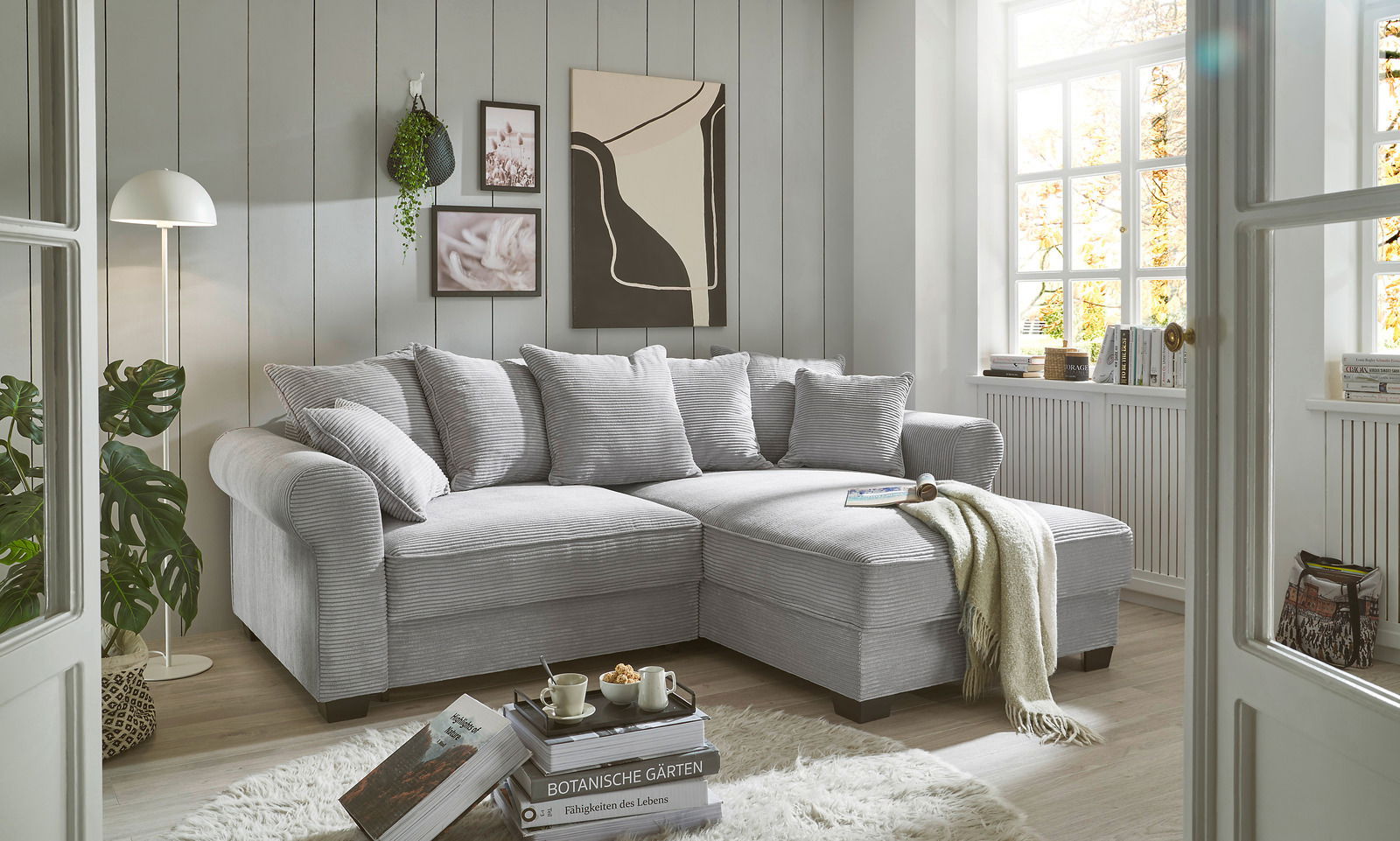 Gemütliche graue Eckcouch in einem hellen Wohnzimmer mit großen Fenstern, dekoriert mit Kissen und einer Decke, seitliche Perspektive.