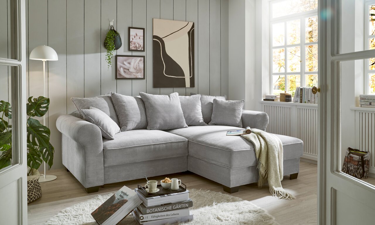 Hom´in Ecksofa MAUREEN Gemütliche graue Eckcouch in einem hellen Wohnzimmer mit großen Fenstern, dekoriert mit Kissen und einer Decke, seitliche Perspektive.