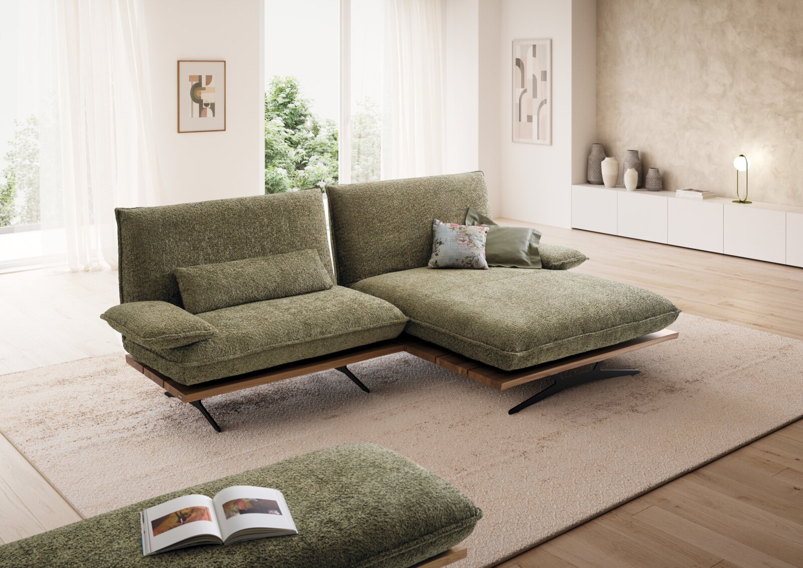 Modernes Ecksofa in olivgrünem Stoff mit breiter Ottomane rechts, Holzsockel und schwarzen Metallfüßen in hellem Wohnzimmer; Perspektive: frontale 3/4-Ansicht von links.