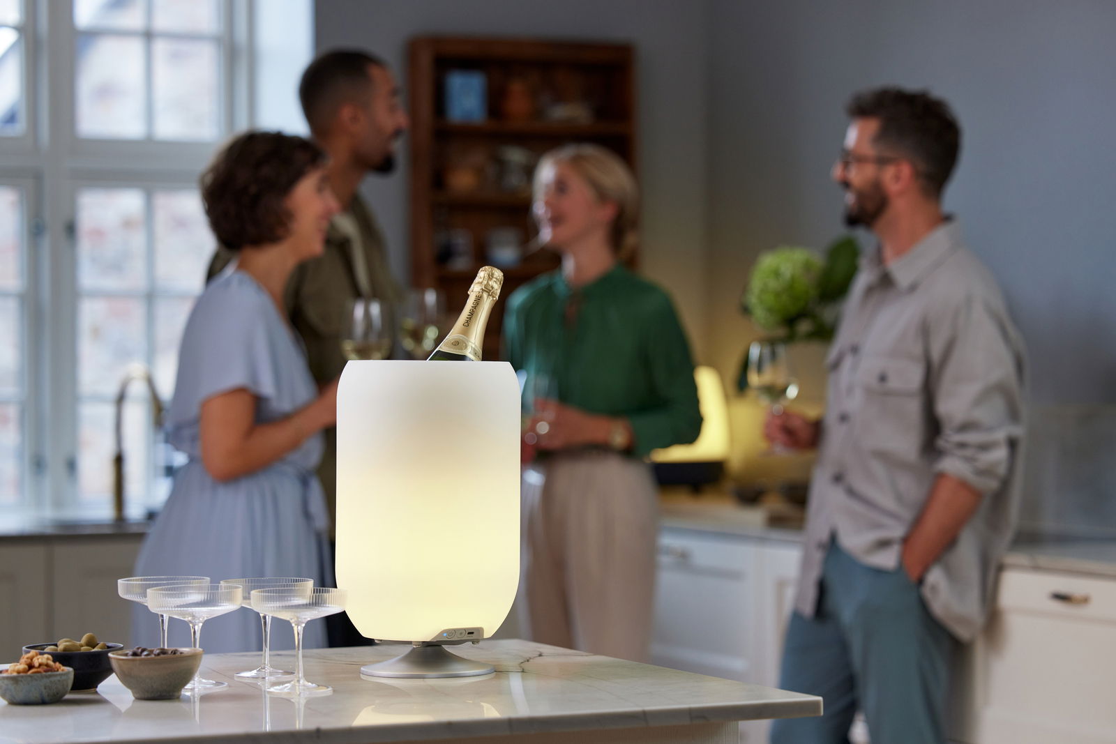 Ein stilvoller, beleuchteter Weinkühler in gebürstetem Silber auf einem Küchentresen, umgeben von vier Personen, die sich unterhalten. Der Weinkühler ist aus der Perspektive von vorne zu sehen.