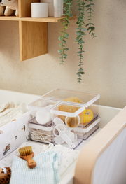 Transparente Aufbewahrungsboxen mit Deckel, gefüllt mit Babyutensilien wie Bürsten und Wattestäbchen, auf einem Wickeltisch von oben betrachtet.