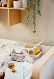 Transparente Aufbewahrungsboxen mit Deckel, gefüllt mit Babyutensilien wie Bürsten und Wattestäbchen, auf einem Wickeltisch von oben betrachtet.