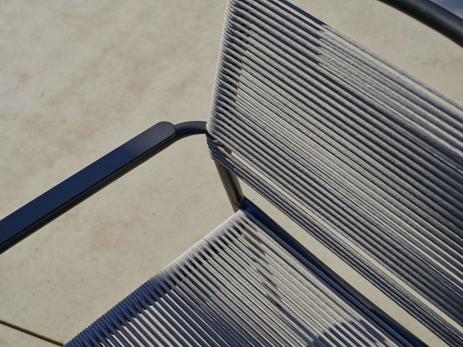 Nahaufnahme eines Stapelsessels mit blauen Armlehnen und grauen Seilbespannungen, von oben betrachtet.