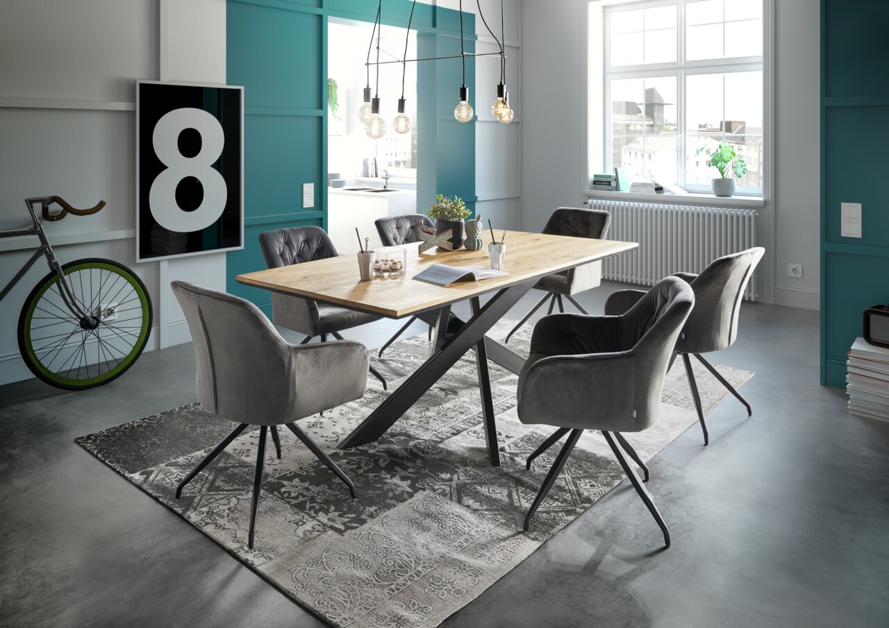 Moderner Esstisch mit Holzplatte und schwarzen Metallbeinen, umgeben von grauen Polsterstühlen in einem stilvoll eingerichteten Raum mit großen Fenstern und dekorativen Elementen. Perspektive: seitliche Ansicht.