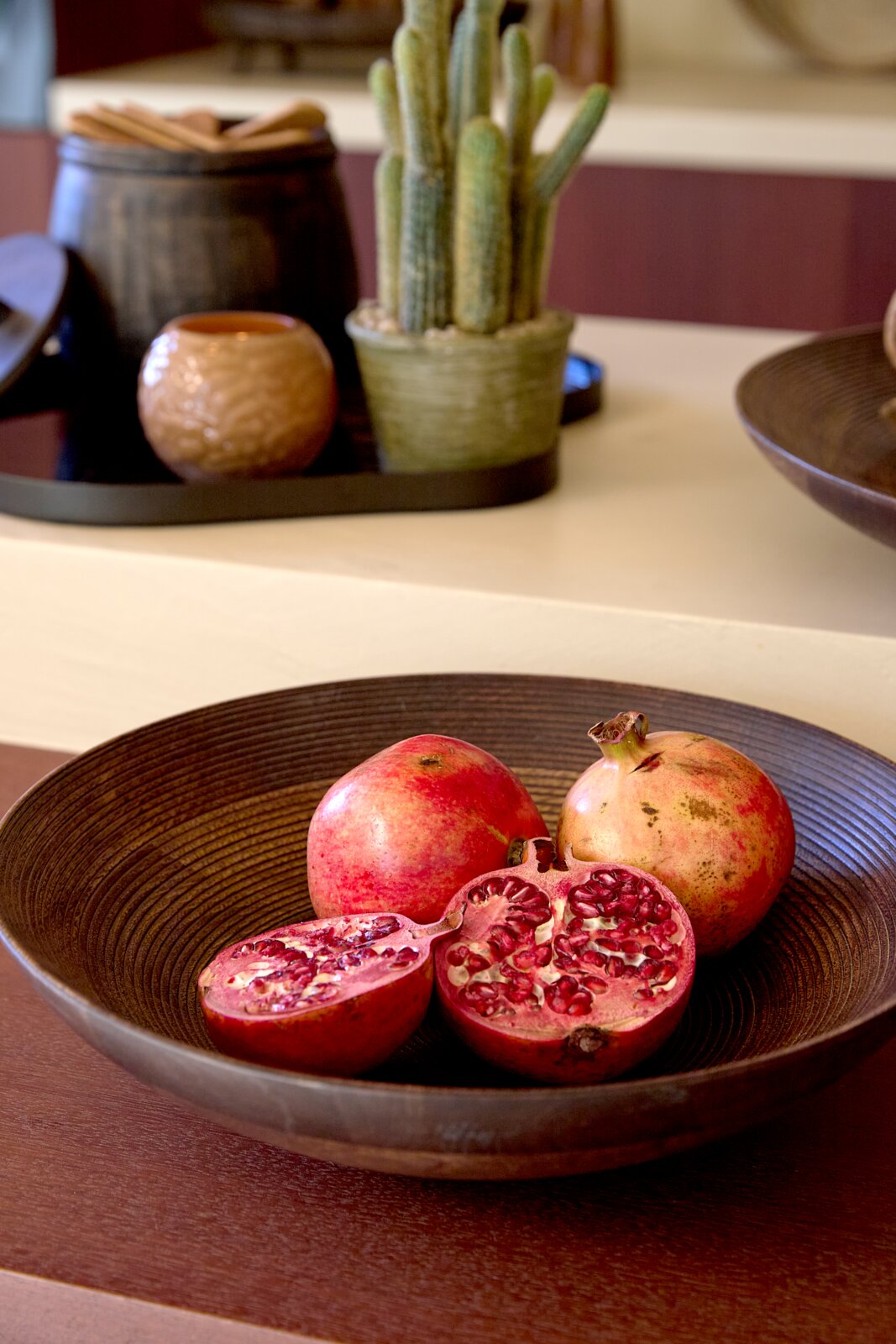 Schuin vooraanzicht van een grote donkerbruine decoratieve schaal met hele en doorgesneden granaatappels op een houten tafel.