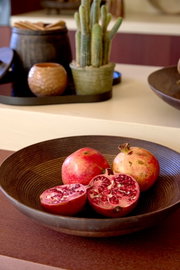 Schuin vooraanzicht van een grote donkerbruine decoratieve schaal met hele en doorgesneden granaatappels op een houten tafel.