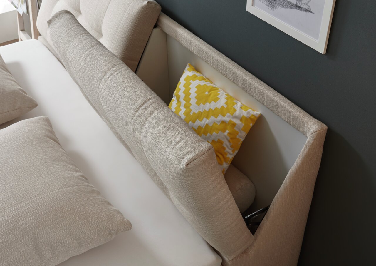 Beige Boxspringbett mit gepolstertem Kopfteil und gelbem Zierkissen, seitliche Perspektive