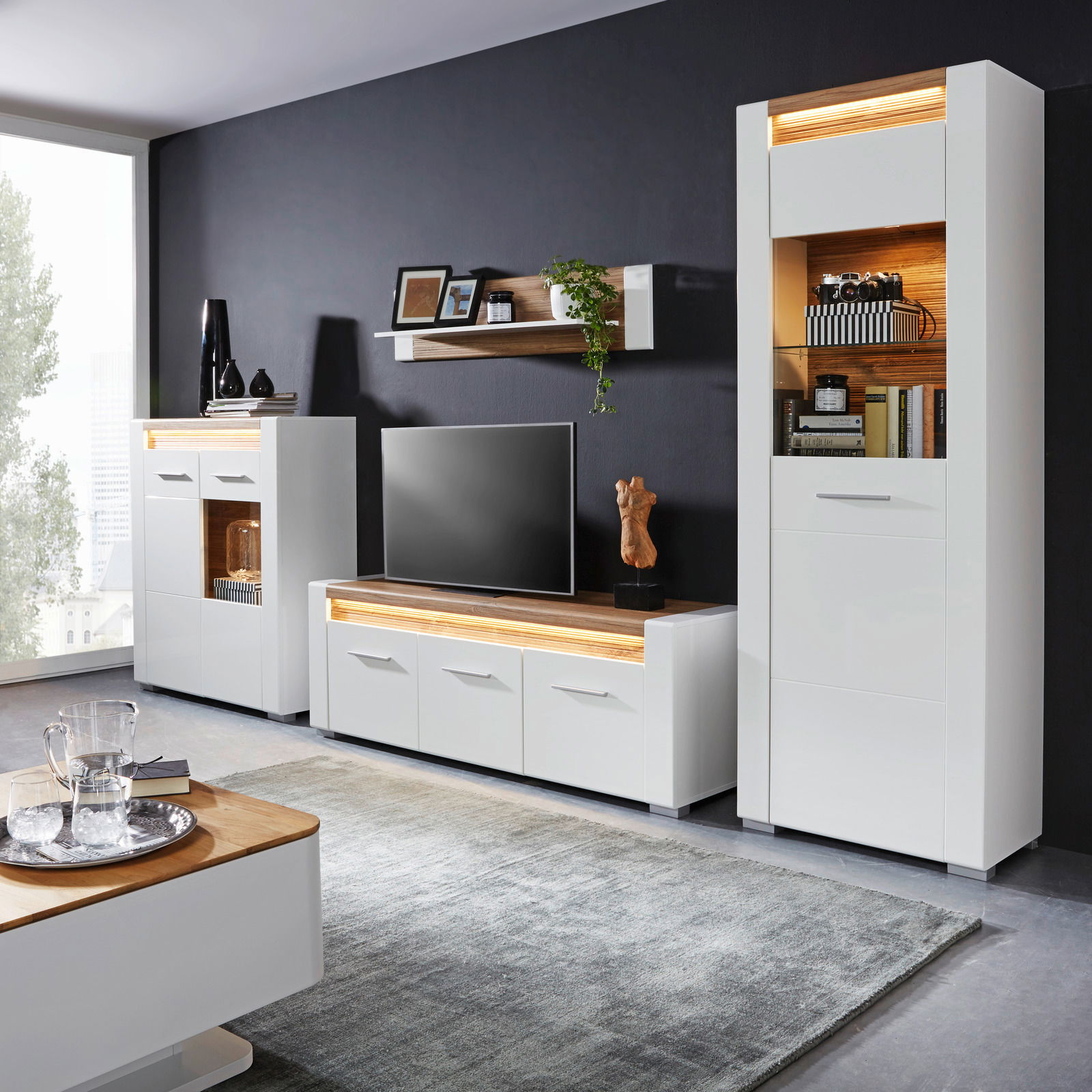 Moderne Wohnzimmermöbel in Weiß mit Holzakzenten, bestehend aus TV-Unterteil, Vitrine und Wandboard, aus der Perspektive des Wohnzimmers betrachtet.