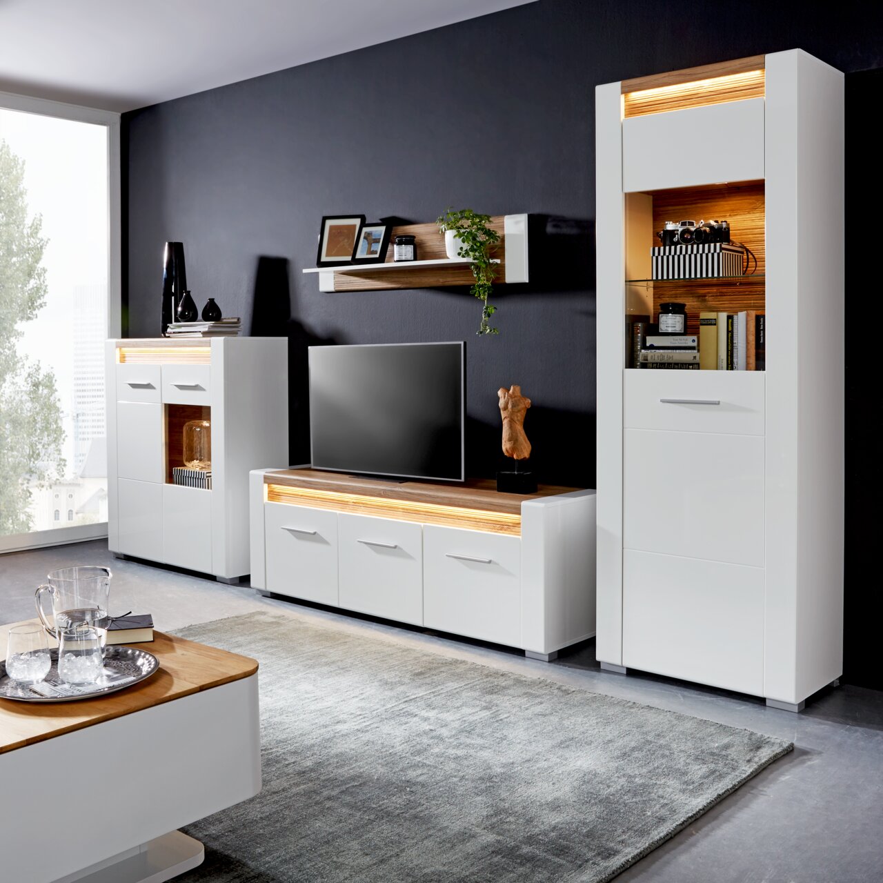 Moderne Wohnzimmermöbel in Weiß mit Holzakzenten, bestehend aus TV-Unterteil, Vitrine und Wandboard, aus der Perspektive des Wohnzimmers betrachtet.