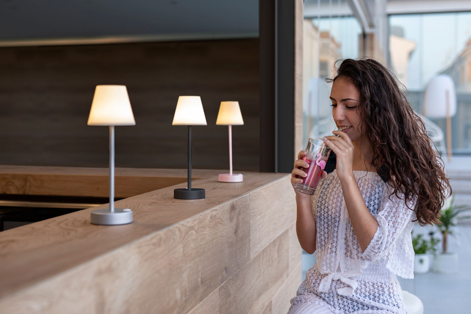 Drei Tischleuchten Lola Slim in verschiedenen Farben auf einer Holztheke, seitliche Perspektive, mit einer Frau, die ein Getränk genießt.
