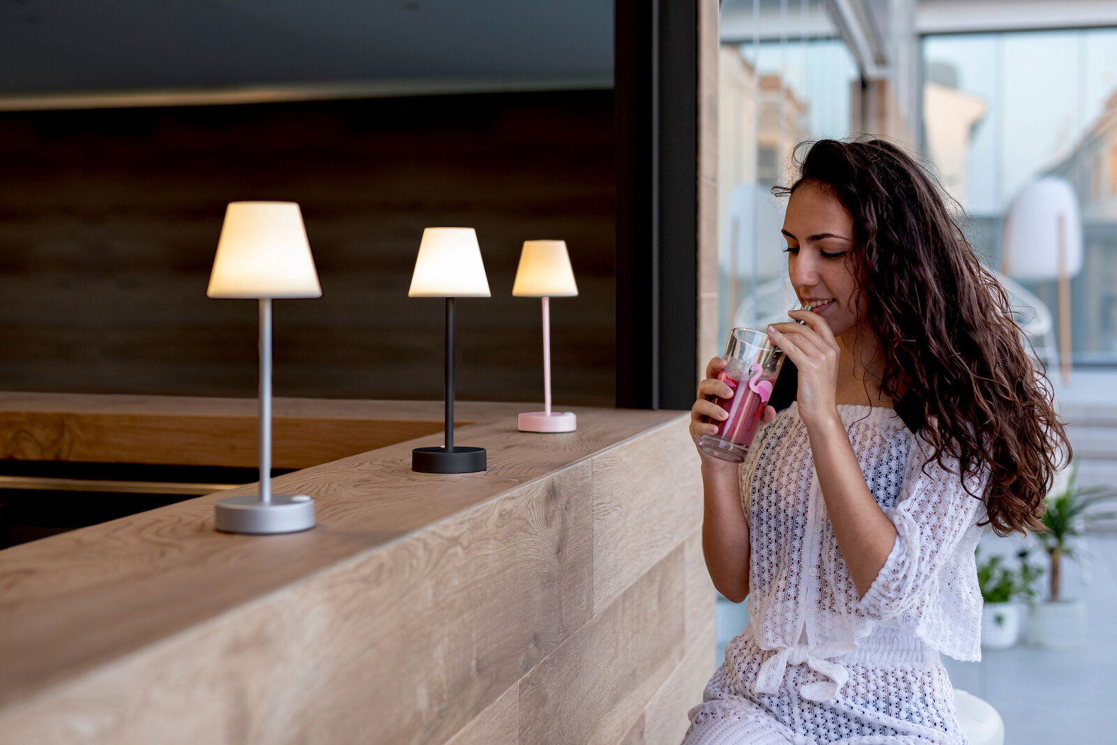 Drei Tischleuchten Lola Slim in verschiedenen Farben auf einer Holztheke, seitliche Perspektive, mit einer Frau, die ein Getränk genießt.