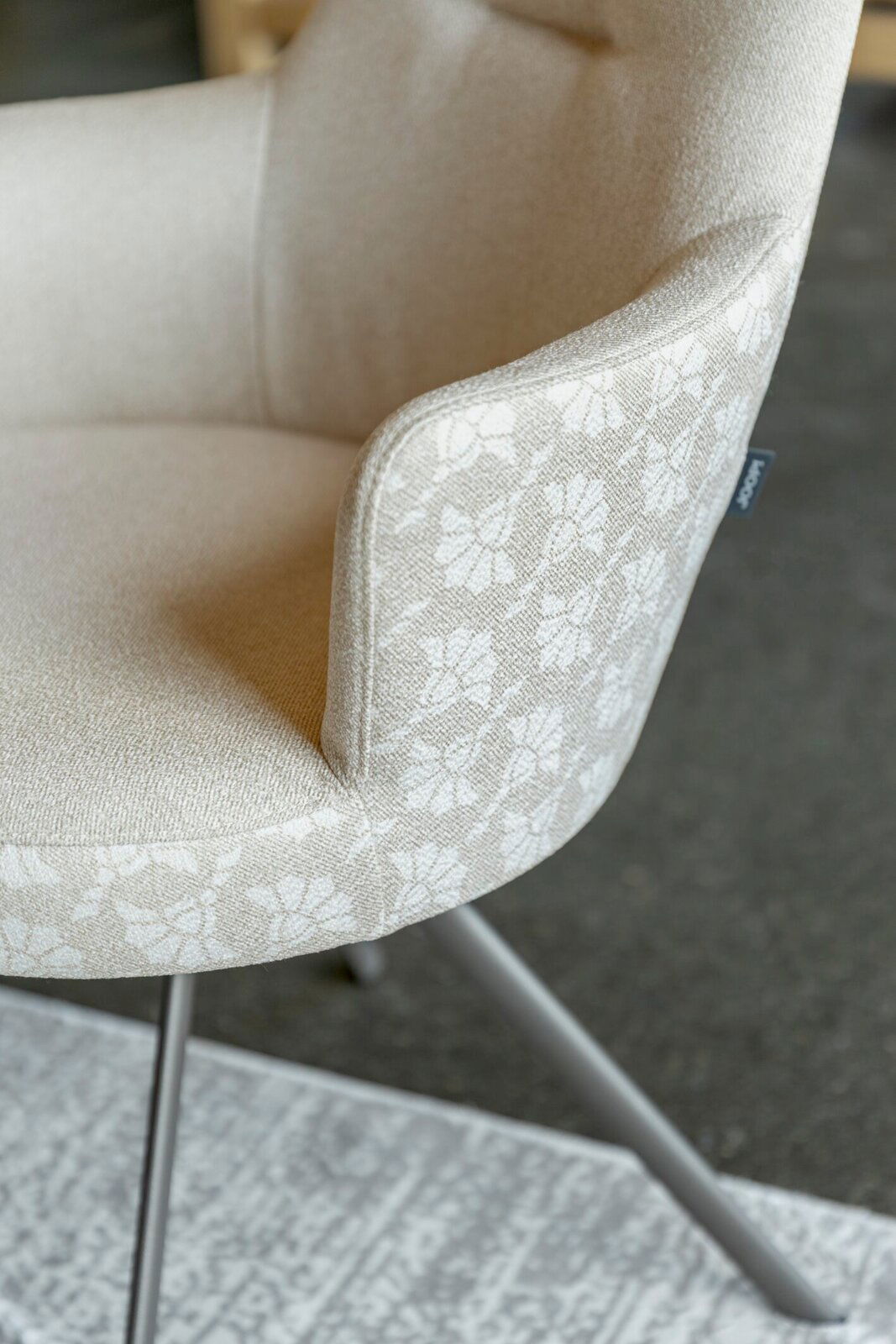 Nahaufnahme eines gepolsterten Stuhls in Beige mit floral gemustertem Stoff an Armlehne und Sitzkante, seitliche Perspektive