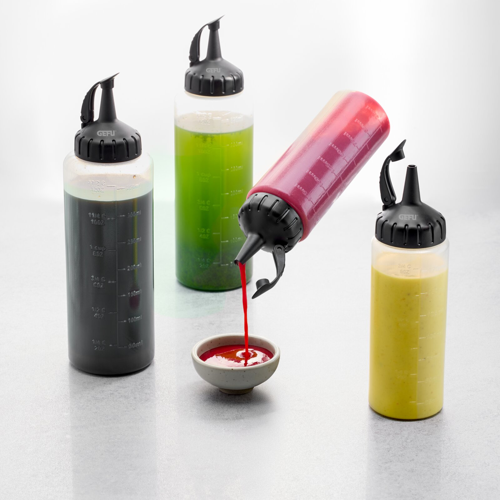 GEFU Dosierflasche FLEXI-FILL Vier Dosierflaschen mit verschiedenen Flüssigkeiten, eine wird schräg gehalten und gießt rote Sauce in eine Schale.