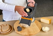 Person reibt Käse mit einer grauen Mandoline/Reibe über einer Glasschüssel auf einem Holzbrett, schräg von oben aufgenommen (Top-Down-Perspektive); daneben Brot, Käseblock und Schüssel mit Käseraspeln.