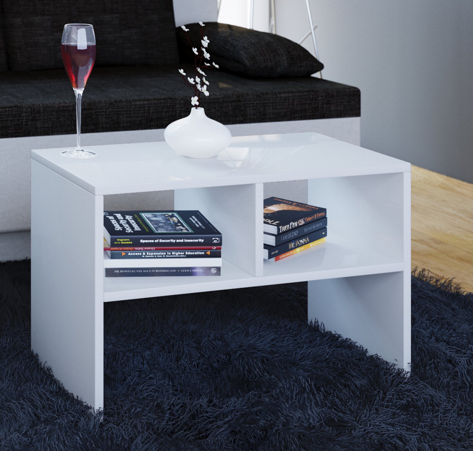 Weißer, moderner Couchtisch mit offener Ablage und Bücherfächern auf dunklem Hochflorteppich vor einem Sofa, mit Vase und Glas auf der Tischplatte, Ansicht schräg von vorne.
