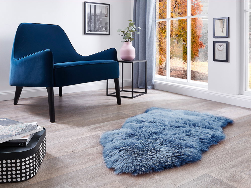 Blauer Samtsessel neben Fenster mit herbstlichem Ausblick, kombiniert mit weichem Fellteppich in kühlen Blautönen und minimalistischer Dekoration
