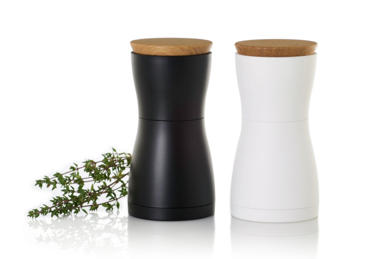 Twee moderne kruidenmolens, een zwarte en een witte, met houten deksels, van voren gefotografeerd, naast een takje verse kruiden.
