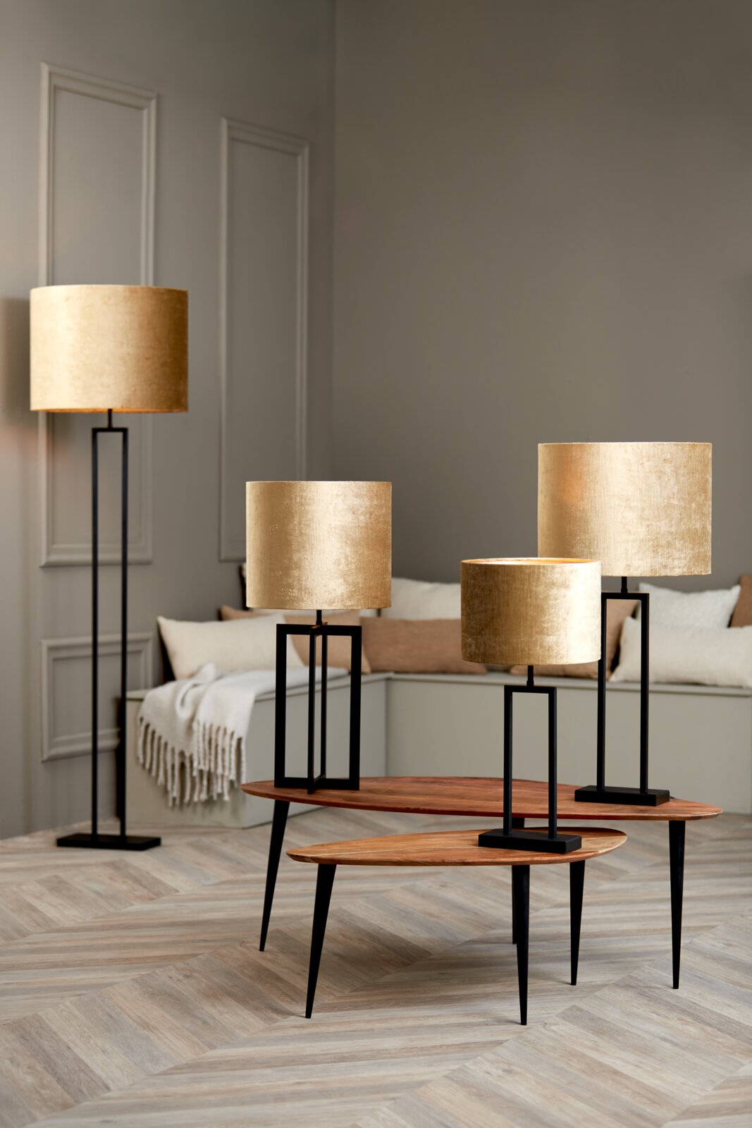 Goldene zylindrische Lampenschirme in verschiedenen Größen auf schwarzen Ständern, fotografiert in einem modernen Wohnzimmer aus der Seitenperspektive.