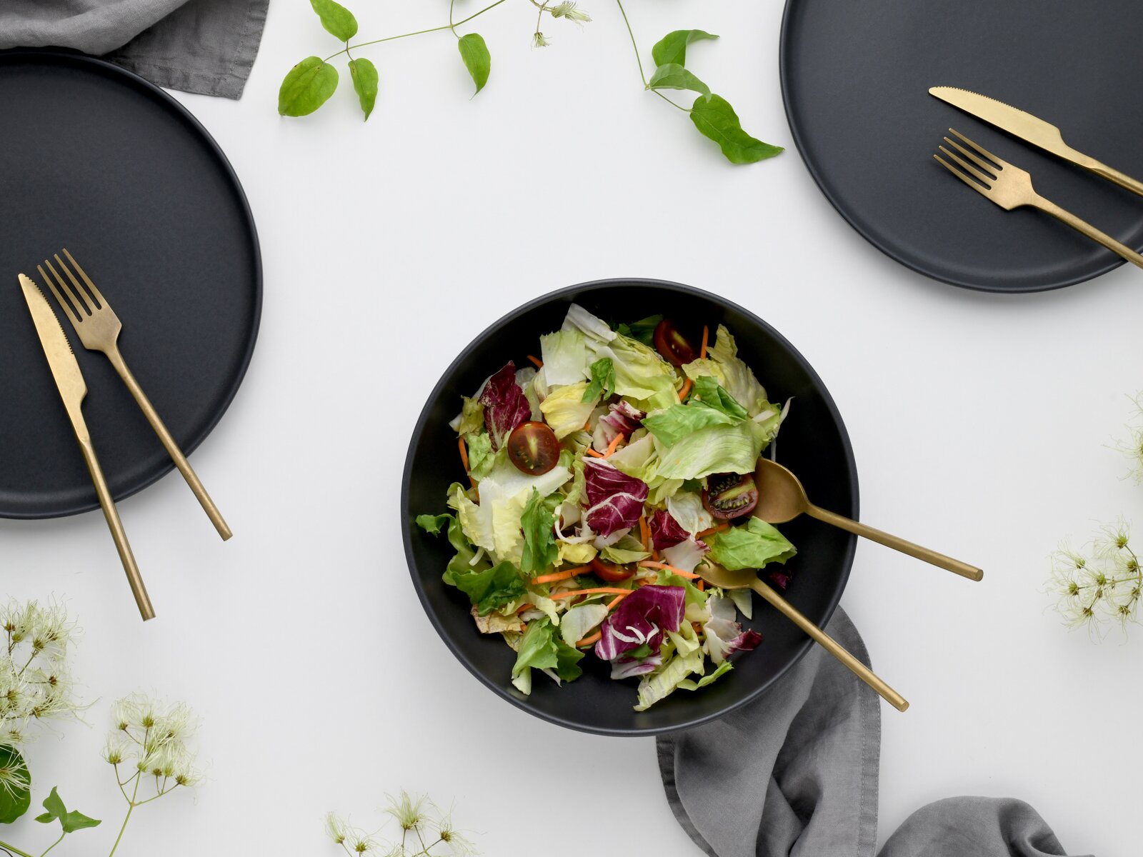 Draufsicht auf einen gedeckten Tisch mit mattschwarzen Tellern und einer schwarzen Schüssel mit buntem Blattsalat, dazu goldfarbenes Besteck und graue Stoffservietten auf weißem Hintergrund.